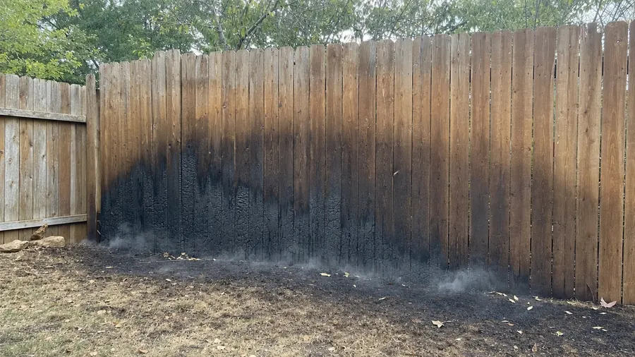 A wide fan-shaped black scorch mark burned across a cedar privacy fence with burned grass in front of it