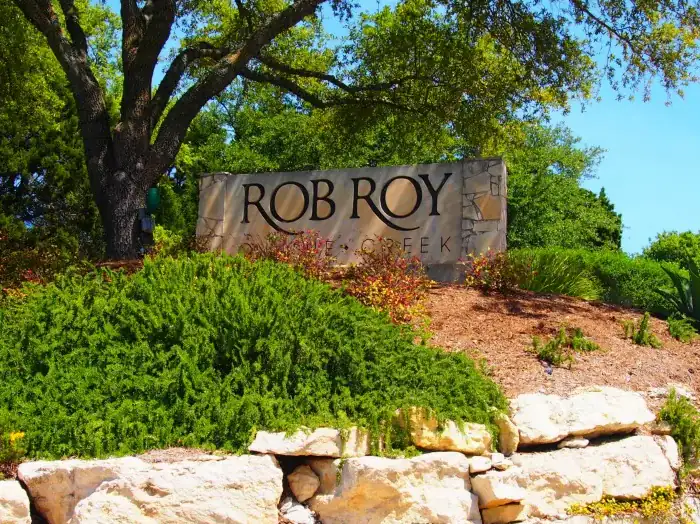 Rob Roy on the Creek limestone entrance sign under a live oak above a boulder wall, West Lake Hills area, Texas