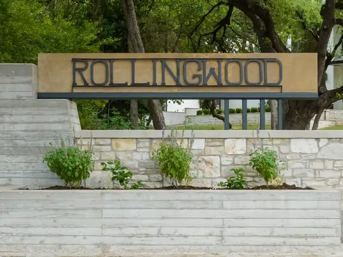 Rollingwood entrance sign on a limestone and board-formed concrete wall under live oaks, near West Lake Hills, Texas